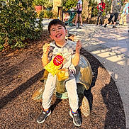 Conrad is registered to the contest to win money with this photo: child, boy, popcorn, popcorn_bag, turtle_statue, statue, sitting, smiling, outdoor, park, stroller, adults, trees, bushes, mulch, sidewalk, sunlight, shadow, sneakers, family