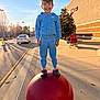 Conrad is registered to the contest to win money with this photo: child, boy, smile, standing, red_bollard, shopping_cart, sidewalk, sunset, sky, hoodie, tracksuit, crocs, shoes, parking_lot, building, tree, shadow, playful, urban, portrait