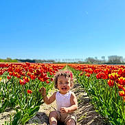 Pharell participe au concours pour gagner de l'argent avec cette photo : toddler, child, flower, tulip, field, outdoor, nature, smiling, happy, sunny, blue_sky, plants, greenery, summer, sitting, casual_clothing, curly_hair, headband, dirt_path, daylight
