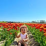toddler, child, flower, tulip, field, outdoor, nature, smiling, happy, sunny, blue_sky, plants, greenery, summer, sitting, casual_clothing, curly_hair, headband, dirt_path, daylight