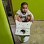 toddler, child, toilet, bathroom, green_wall, floor, tiles, sitting, serious_expression, buzz_cut, white_toilet, indoor, plumbing_fixture, person, casual_clothing, short_sleeves, holding, concrete_floor, wall_damage, curious