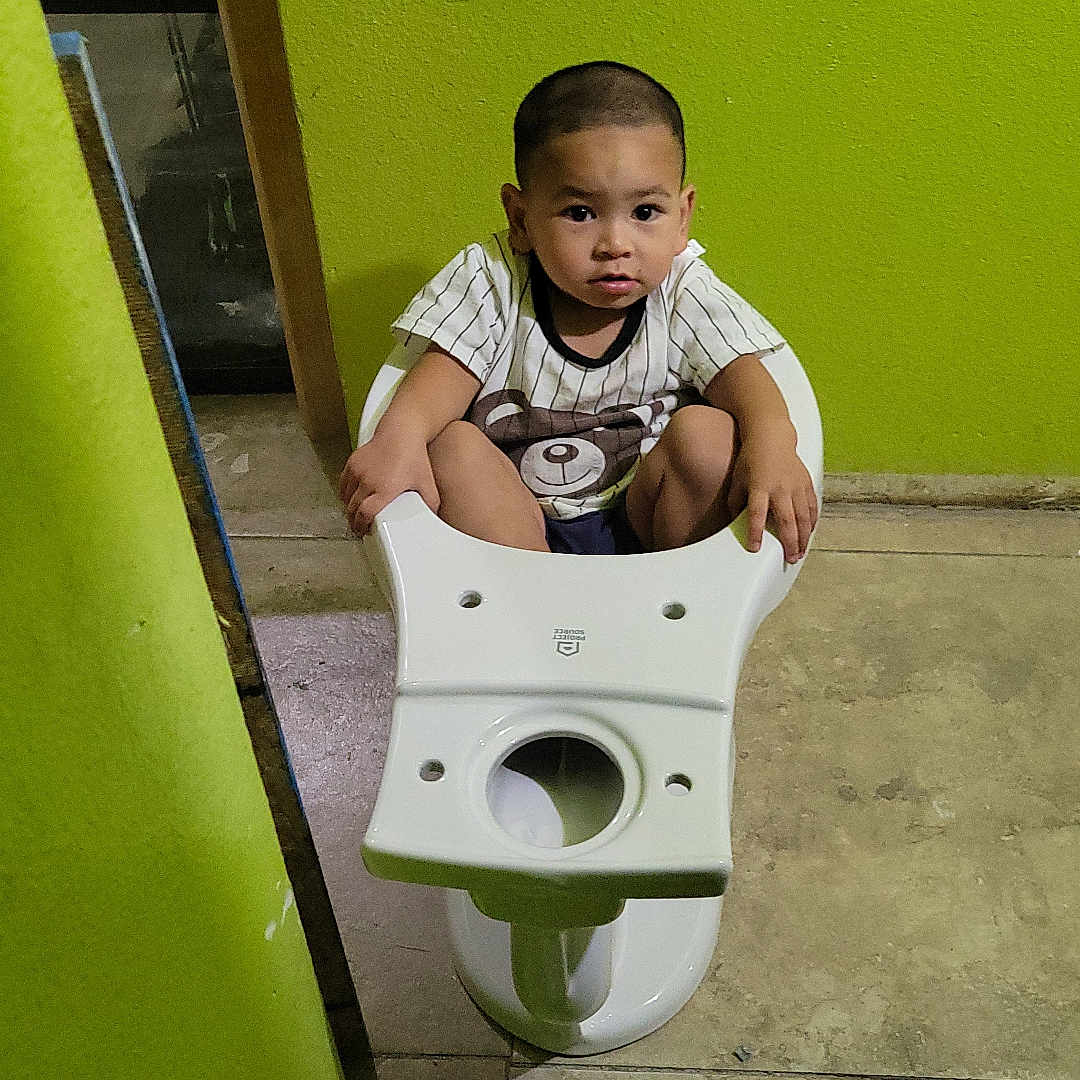 Ozel joined the competition — help win amazing prizes! bathroom, buzz_cut, casual_clothing, child, concrete_floor, curious, floor, green_wall, holding, indoor, person, plumbing_fixture, serious_expression, short_sleeves, sitting, tiles, toddler, toilet, wall_damage, white_toilet