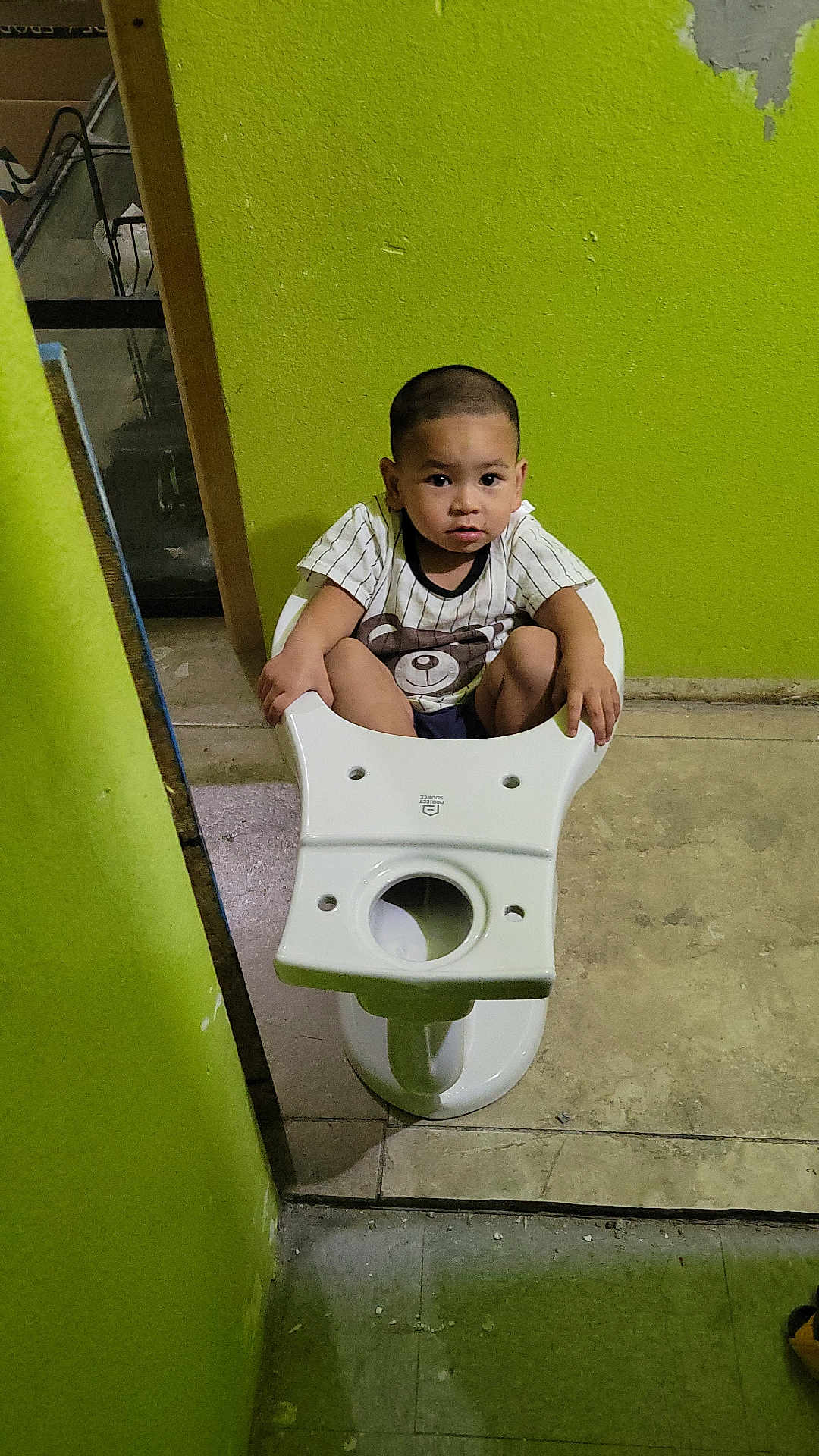 Ozel joined the competition — help win amazing prizes! toddler, child, toilet, bathroom, green_wall, floor, tiles, sitting, serious_expression, buzz_cut, white_toilet, indoor, plumbing_fixture, person, casual_clothing, short_sleeves, holding, concrete_floor, wall_damage, curious