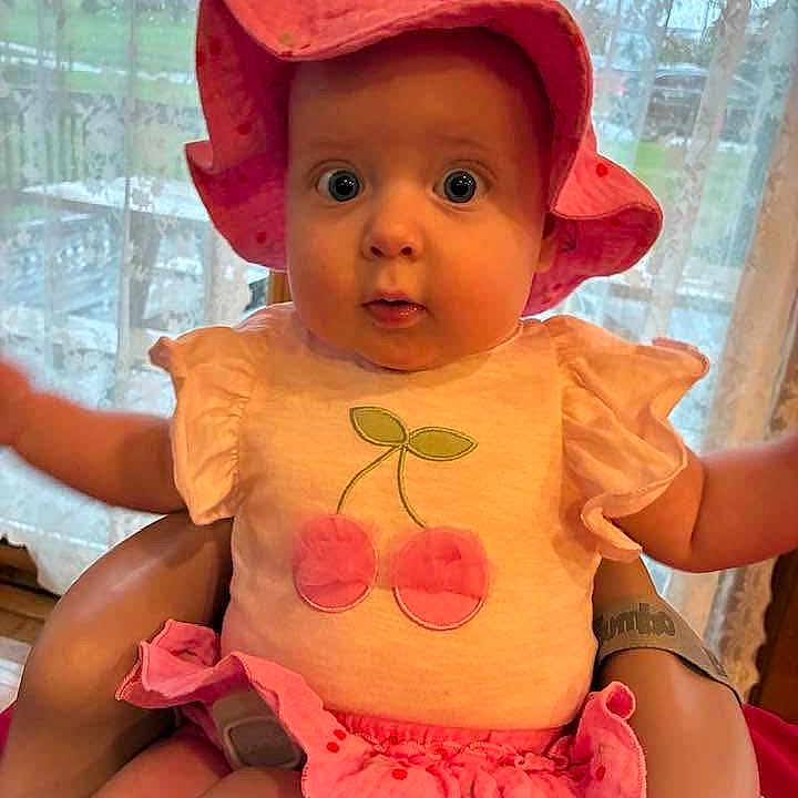 Sabrina is registered to the contest to win money with this photo: adorable, arms_outstretched, baby, cherry_design, child, clothing, curtains, cute, expression, holding, indoor, infant, person, pink_hat, pink_skirt, portrait, sitting, soft_light, wide_eyes, window