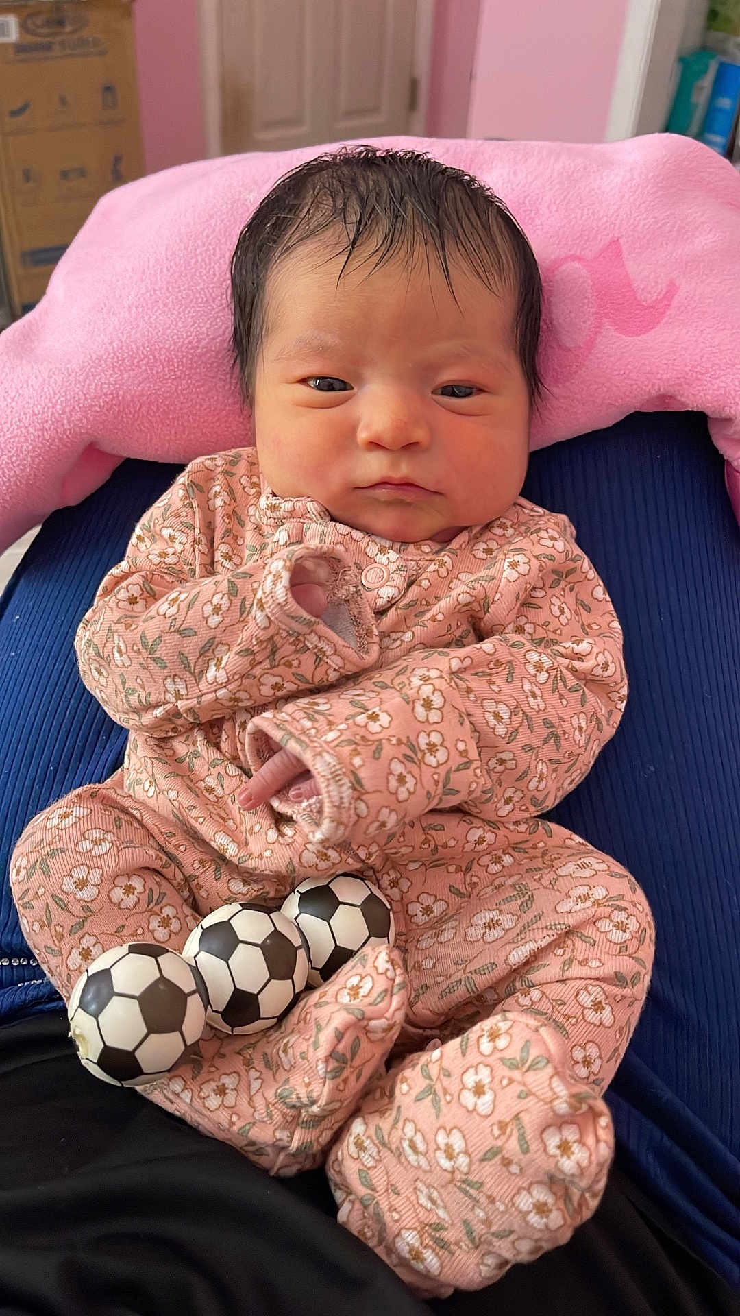 Aurora is registered to the contest to win money with this photo: newborn, baby, infant, onesie, pink, floral_pattern, toy, soccer_balls, cushion, pillow, indoor, person, face, head, clothing, sleepwear, cute, resting, child, small