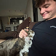 Tulo is registered to the contest to win money with this photo: cat, person, smiling, indoor, couch, petting, tabby_cat, black_shirt, modern_kitchen, furniture, cozy, relaxed, home, cushion, collar, brown_couch, short_hair, happy, domestic_animal, casual