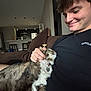 cat, person, smiling, indoor, couch, petting, tabby_cat, black_shirt, modern_kitchen, furniture, cozy, relaxed, home, cushion, collar, brown_couch, short_hair, happy, domestic_animal, casual