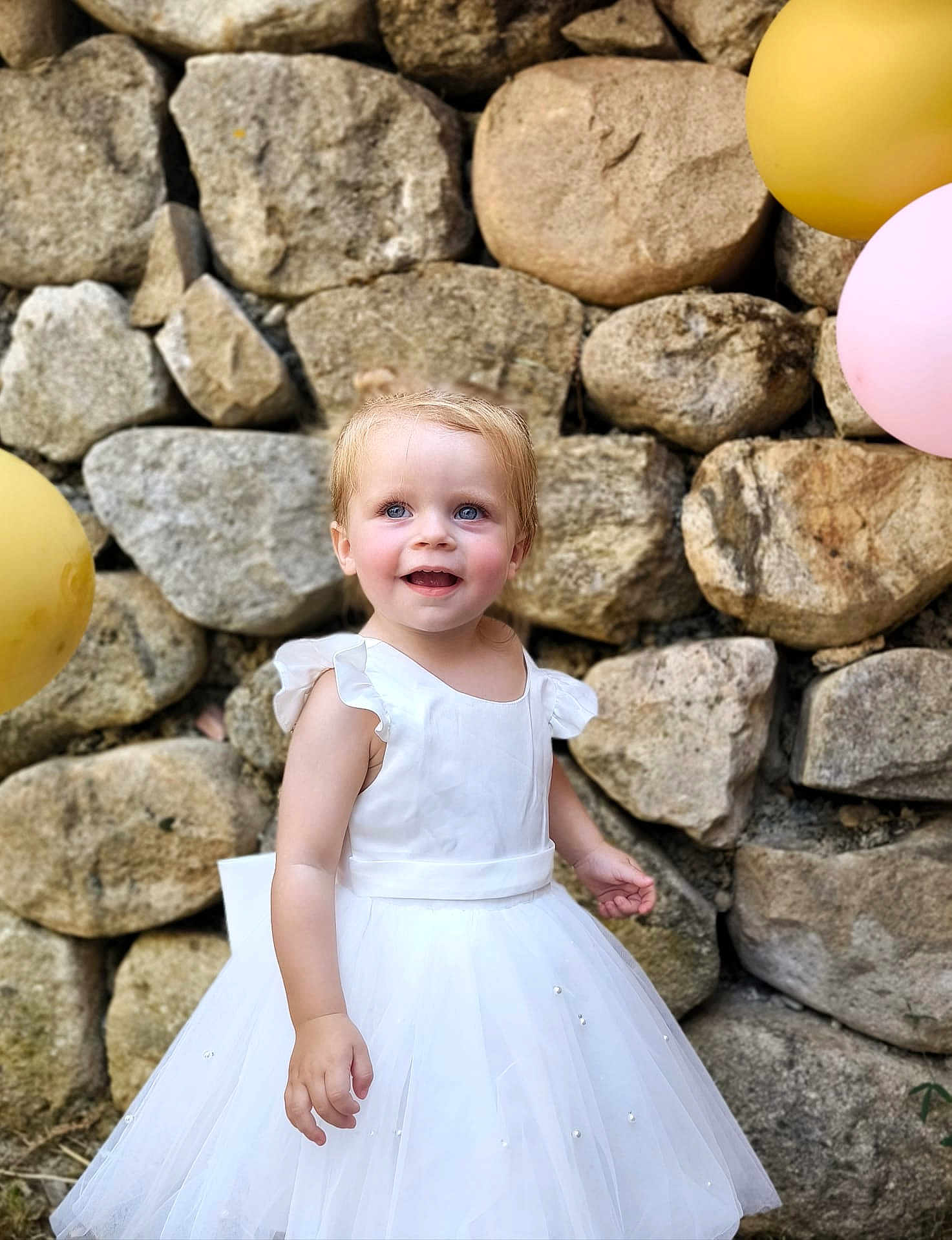 Elise participe au concours pour gagner de l'argent avec cette photo : toddler, child, white_dress, balloons, stone_wall, smiling, outdoor, happy, portrait, cute, celebration, party, young_child, person, fashion, dress, event, colorful, fun, joyful