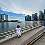 child, water, sky, cityscape, buildings, railing, clothing, white_outfit, curly_hair, urban, outdoor, side_view, standing, stone_ledge, daylight, clouds, river, person, back_view, footwear