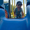 child, toddler, playground, slide, blue_slide, cap, nike, overalls, smiling, outdoor, yellow_bars, sky, trees, play, fun, happy, clothing, person, daytime, recreation