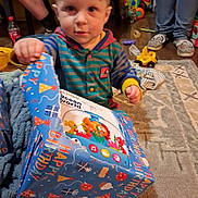 Ryder joined the competition — help win amazing prizes! toddler, child, birthday, present, gift_wrapping, toys, striped_hoodie, indoor, carpet, floor, person, unwrapping, celebration, curious, young_child, home, colorful, playtime, happy, living_room