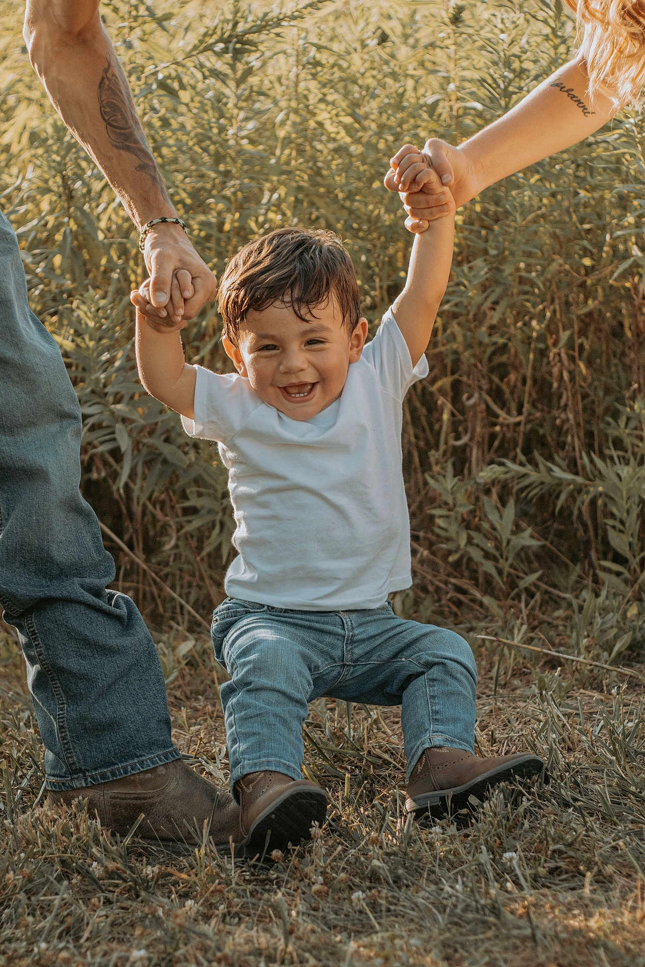 Giovanni is registered to the contest to win money with this photo: child, toddler, smiling, happy, white_tshirt, jeans, boots, grass, outdoor, nature, holding_hands, tattoo, adult, sunlight, greenery, casual_clothing, family, playful, portrait, joy
