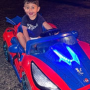 Giovanni joined the competition — help win amazing prizes! child, toddler, toy_car, red, blue, night, outdoor, smiling, happy, steering_wheel, vehicle, plastic, driveway, casual_clothing, shorts, tshirt, headlight, wheel, car_toy, person