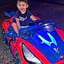 child, toddler, toy_car, red, blue, night, outdoor, smiling, happy, steering_wheel, vehicle, plastic, driveway, casual_clothing, shorts, tshirt, headlight, wheel, car_toy, person
