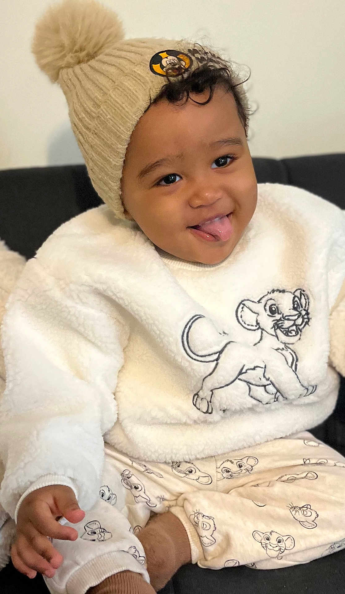 Lucas participe au concours pour gagner de l'argent avec cette photo : child, baby, toddler, face, smile, tongue_out, curly_hair, beanie, pompom_hat, sweater, lion_king, simba, patterned_pants, cozy, plush_texture, hand, seated, couch, indoors, happy