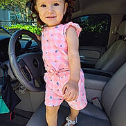 Isabel Escobedo is registered to the contest to win money with this photo: car_interior, child, curly_hair, cute, dashboard, footwear, greenery, hair_clips, hand, house, pink_clothing, playful, portrait, sandals, seat, smiling, steering_wheel, sunlight, toddler, window