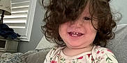 Isabel Escobedo joined the competition — help win amazing prizes! toddler, child, curly_hair, smiling, indoor, couch, pillow, floral_shirt, home_decor, window_shutters, lamp, painting, happy, person, face, seated, casual_clothing, interior, cute, portrait