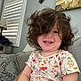 toddler, child, curly_hair, smiling, indoor, couch, pillow, floral_shirt, home_decor, window_shutters, lamp, painting, happy, person, face, seated, casual_clothing, interior, cute, portrait