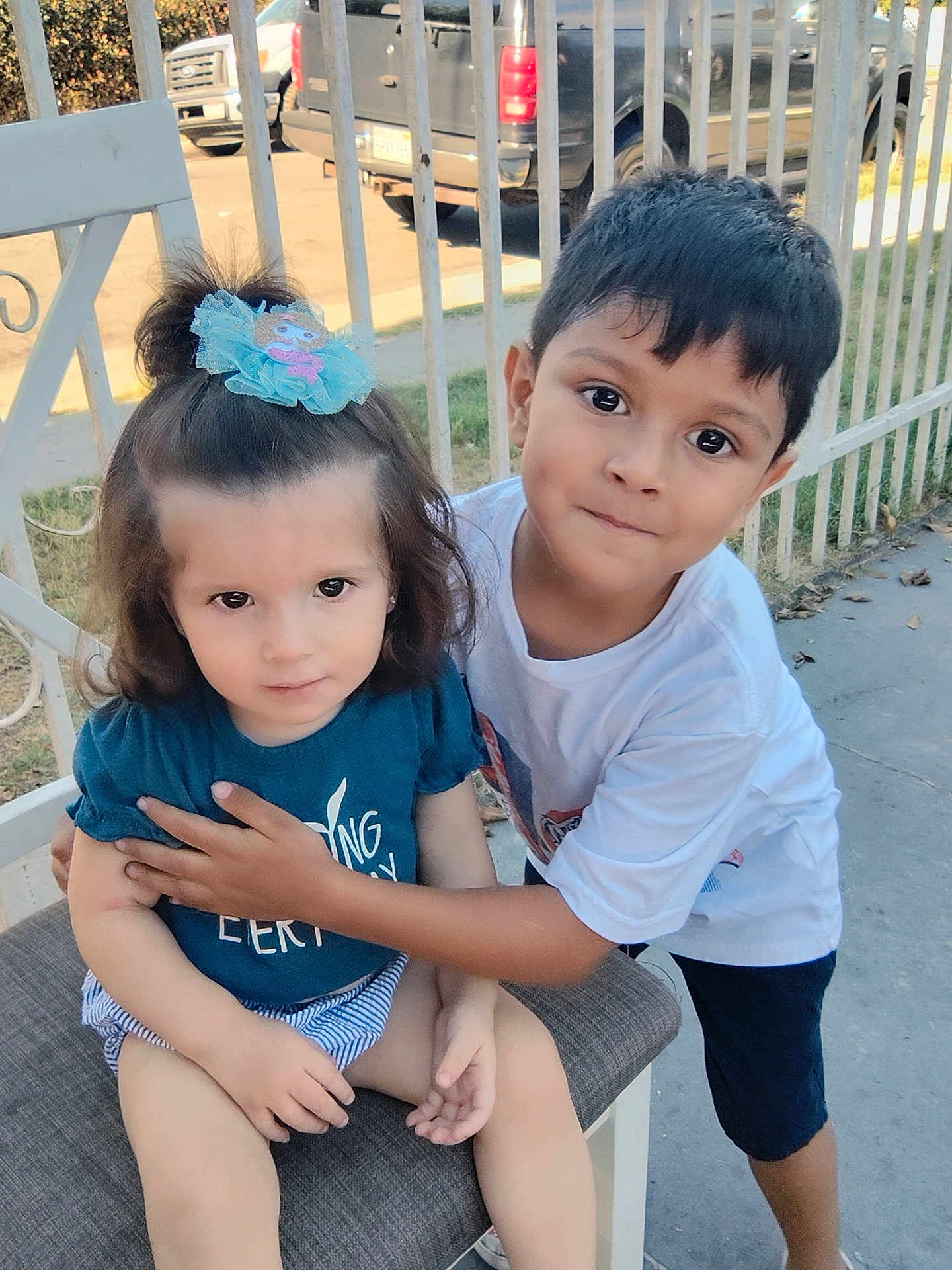 Gael Escobedo is registered to the contest to win money with this photo: child, children, boy, girl, outdoor, fence, chair, hair_accessory, shorts, tshirt, sitting, hugging, curious, daylight, concrete, smiling, parked_vehicle, playful, young, affection