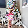 child, toddler, pink_clothing, tutu, earmuffs, unicorn, plush_toy, christmas_tree, pastel_colors, ornaments, holiday_decor, reindeer, owl, nutcracker, wooden_floor, indoor, smiling, festive, cute, barefoot