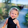 child, blonde_hair, blue_eyes, outdoor, sunny_day, black_top, sleeveless, curly_hair, young_child, nature, pathway, greenery, portrait, expression, holding_object, casual_clothing, daylight, skin, person, cute