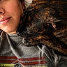 affection, brindle_dog, casual_clothing, close_up, colorful_shirt, comfort, companion, dog, expression, face, human_animal_bond, indoor, lying_down, person, portrait, relaxation, resting, snuggling, soft_lighting, warmth