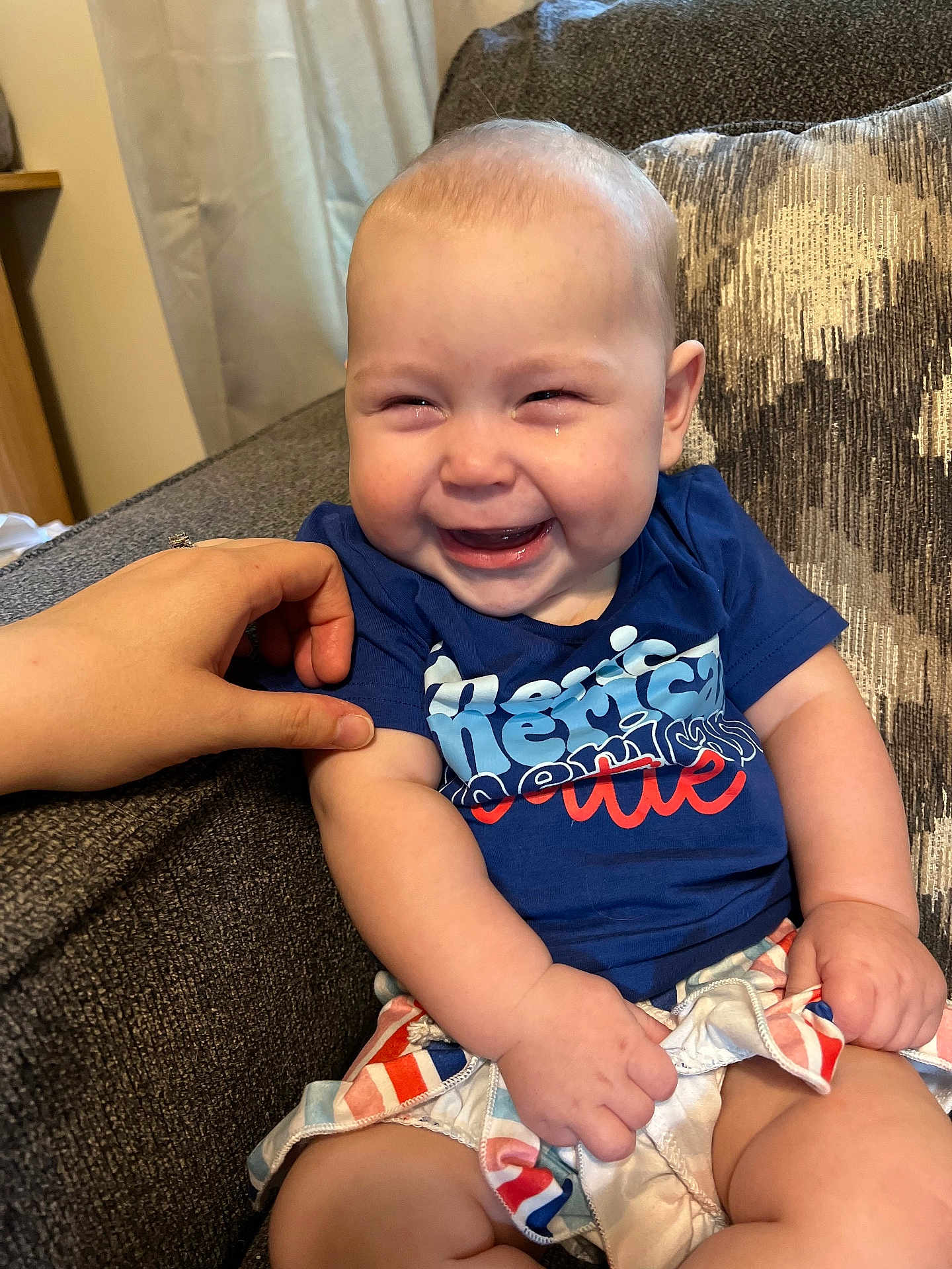 Sophia is registered to the contest to win money with this photo: baby, smiling, laughing, child, blue_shirt, colorful_shorts, couch, hand, tickling, happy, indoor, curtain, fabric, person, skin, face, chubby_arms, cute, comfort, seated