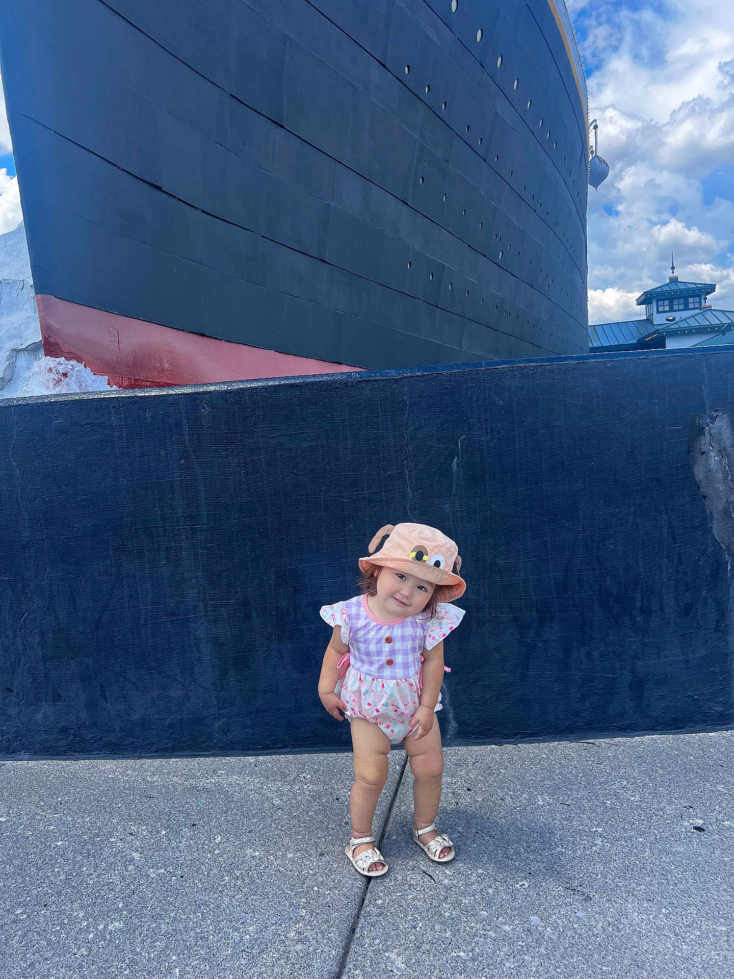 Alora joined the competition — help win amazing prizes! toddler, child, hat, bucket_hat, romper, sandals, concrete, ship, large_vessel, blue_sky, clouds, outdoor, standing, cute, playful, smiling, head_tilt, summer_clothing, daytime, background