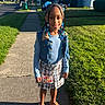 child, girl, smiling, portrait, outdoor, sidewalk, neighborhood, residential, sunlight, grass, houses, denim, plaid, skirt, sandals, braids, hair_beads, blue_clothing, fashion, standing