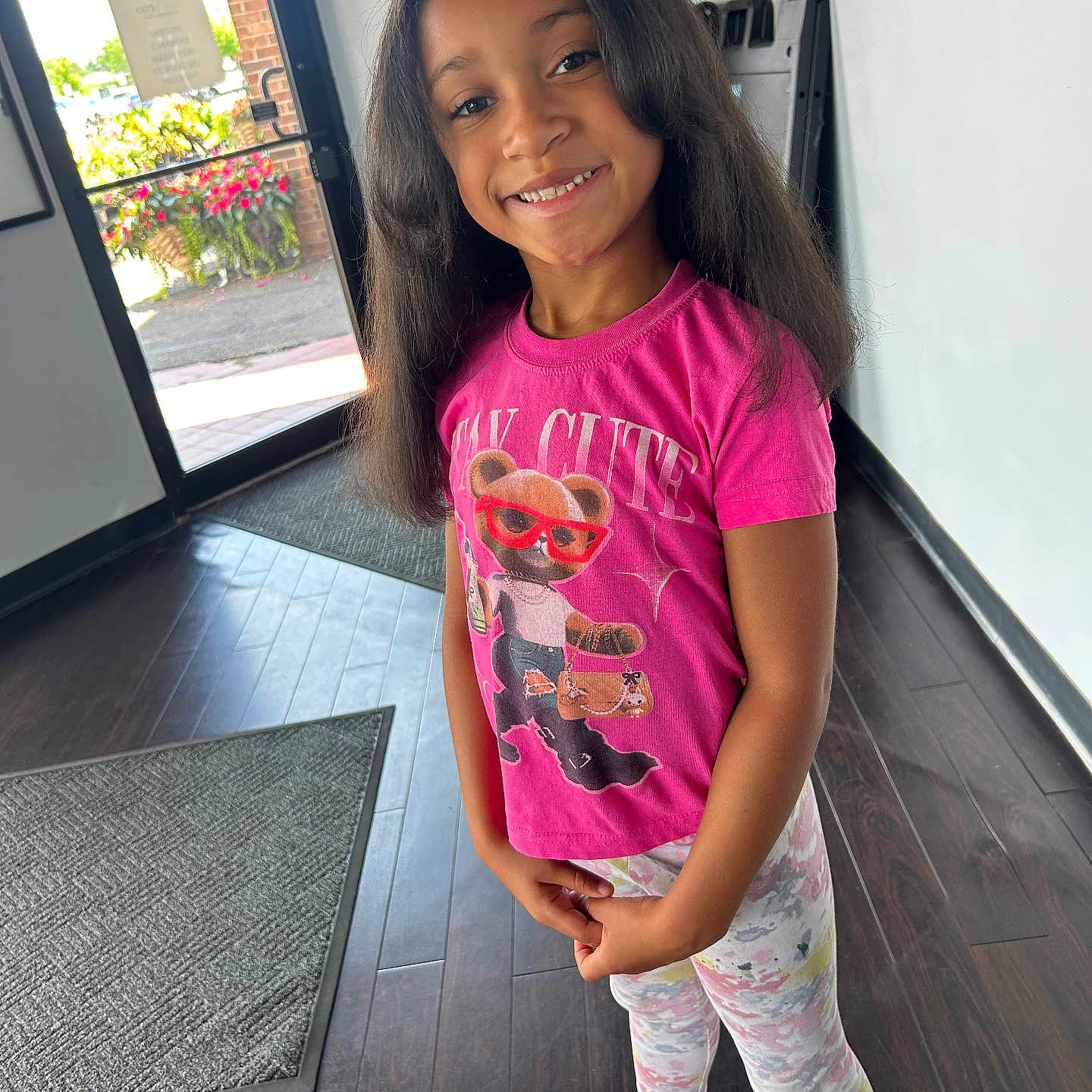 Khrystlyn is registered to the contest to win money with this photo: child, girl, smiling, portrait, standing, indoors, doorway, entrance, pink_shirt, graphic_t_shirt, leggings, pink_shoes, clogs, long_hair, dark_hair, happy, floor, doormat, flowers, daylight