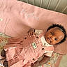 Peyton is registered to the contest to win money with this photo: baby, pink_dress, blanket, embroidery, face, child, infant, curly_hair, lying_down, quilt, pattern, cute, soft_lighting, indoors, hand, footwear, fabric, portrait, skin, clothing