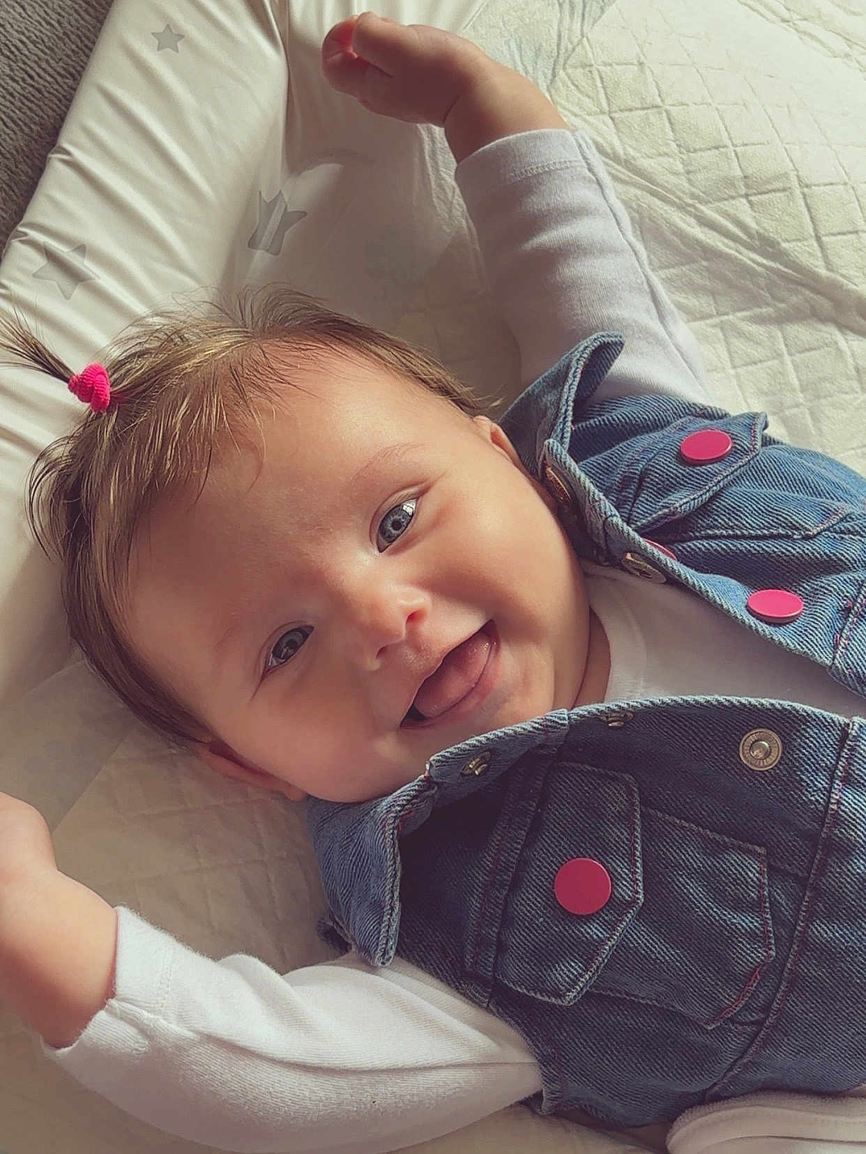 Ariella is registered to the contest to win money with this photo: baby, smiling, denim_vest, pink_hair_tie, lying_down, cute, child, infant, happy, face, blue_eyes, white_shirt, soft_cushion, star_pattern, portrait, closeup, indoor, skin, young_child, joyful