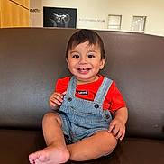 Elijah is registered to the contest to win money with this photo: toddler, child, smiling, sitting, overalls, red_shirt, leather_couch, indoor, portrait, happy, baby_photo, background_wall, short_hair, cute, person, furniture, casual_clothing, feet, bright, young_child