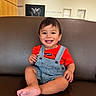toddler, child, smiling, sitting, overalls, red_shirt, leather_couch, indoor, portrait, happy, baby_photo, background_wall, short_hair, cute, person, furniture, casual_clothing, feet, bright, young_child