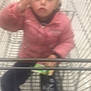 child, shopping_cart, pink_jacket, blurry, indoor, holding_object, young_child, casual_clothing, shopping, person, legs, footwear, metal_cart, store, toy_or_snack, hand, face, expressionless, winter_clothing, small_child