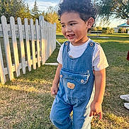 Kaden is registered to the contest to win money with this photo: child, toddler, curly_hair, smiling, outdoor, backyard, grass, white_fence, denim_overalls, t_shirt, sneakers, portrait, standing, sunset, golden_hour, playground, trees, sky, park, happy