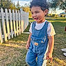 child, toddler, curly_hair, smiling, outdoor, backyard, grass, white_fence, denim_overalls, t_shirt, sneakers, portrait, standing, sunset, golden_hour, playground, trees, sky, park, happy