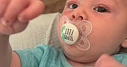 Johnny is registered to the contest to win money with this photo: baby, infant, pacifier, blue_shirt, face, hand, couch, patterned_cushion, closeup, curious, indoors, child, person, young_child, sleeve, text_on_clothing, skin, head, eyes, mouth