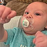 baby, infant, pacifier, blue_shirt, face, hand, couch, patterned_cushion, closeup, curious, indoors, child, person, young_child, sleeve, text_on_clothing, skin, head, eyes, mouth