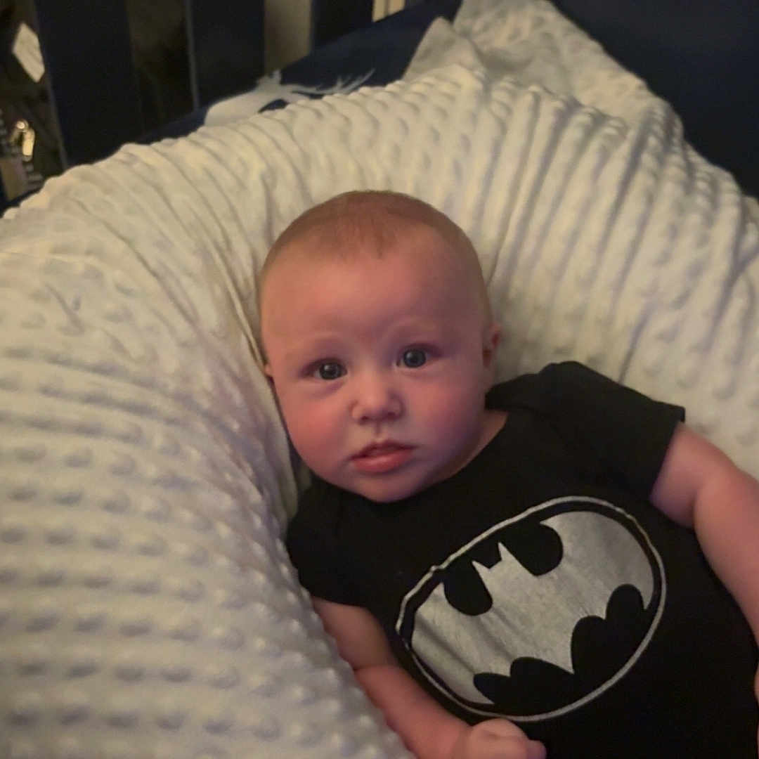 Johnny joined the competition — help win amazing prizes! baby, infant, onesie, batman_logo, pillow, crib, baby_clothing, white_pillow, black_onesie, baby_boy, infant_face, baby_lying_down, baby_bed, baby_portrait, baby_skin, baby_hair, indoor, child, cute, baby_hand