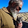 man, beard, sunglasses, cat, tabby_cat, holding, outdoor, desert, hills, sky, sunlight, portrait, nature, scenic, animal, pet, clothing, hand, closeup, casual