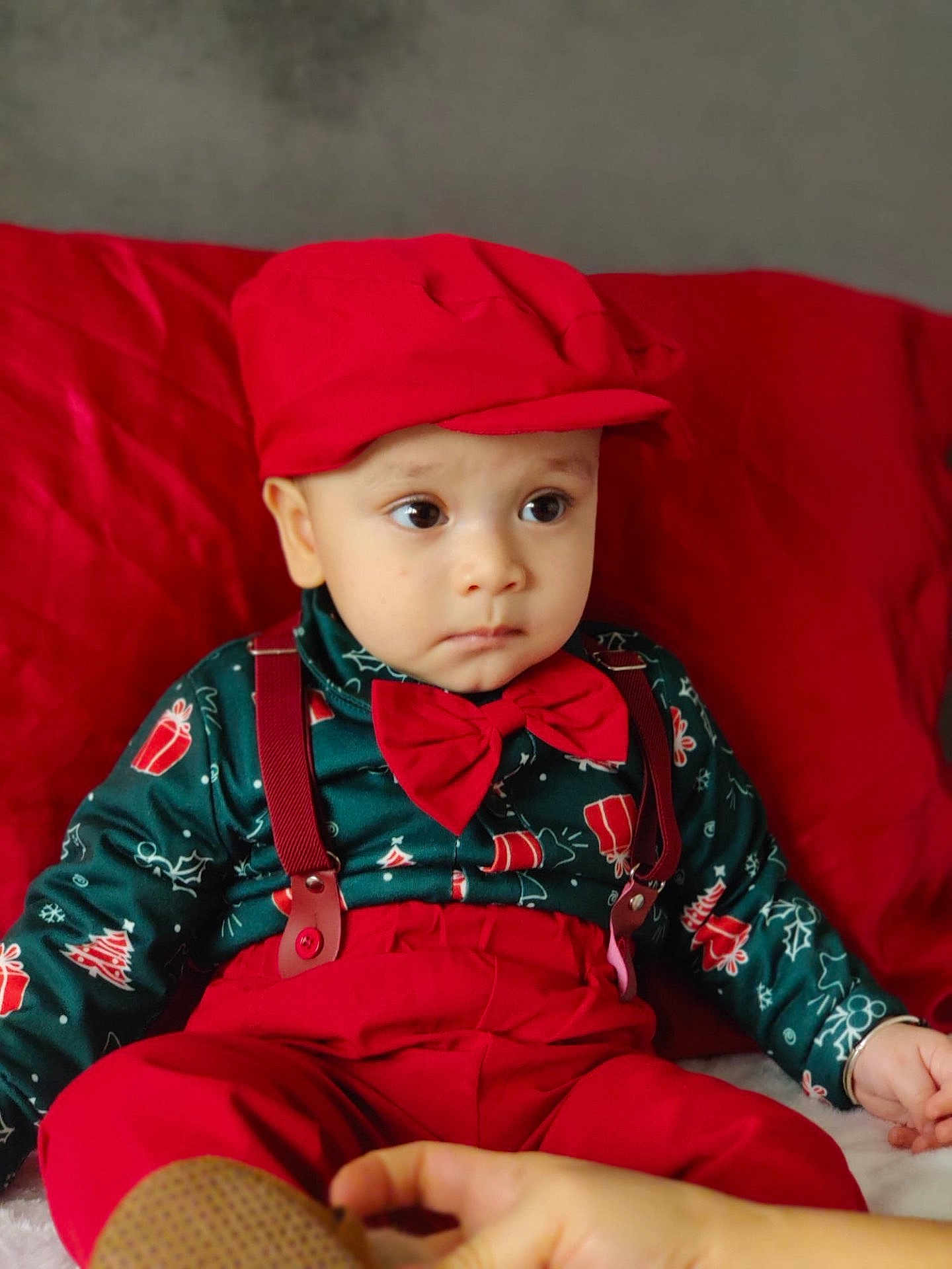 Yuvin is registered to the contest to win money with this photo: baby, child, toddler, red_clothing, bow_tie, suspenders, hat, face, person, indoor, cushion, red_cushion, sitting, cute, expression, hand, shoe, holiday_theme, patterned_shirt, portrait