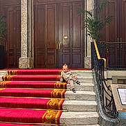 Rafaël participe au concours pour gagner de l'argent avec cette photo : child, stairs, carpet, marble, wooden_doors, columns, plant, railing, interior, luxury, architecture, casual_pose, red, gold, elegant, young, person, fashion, relaxing, indoor