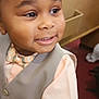 child, toddler, smile, bow_tie, vest, peach_shirt, formal_wear, closeup, portrait, indoor, church_pew, wooden_bench, curly_hair, eyes, skin, buttons, cheek, happy, young_child, floor_carpet