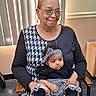 elderly_woman, baby, indoor, smile, woman, child, seated, headband, glasses, patterned_clothing, black_and_white, affection, holding, window_blinds, chair, nail_polish, portrait, person, family, casual
