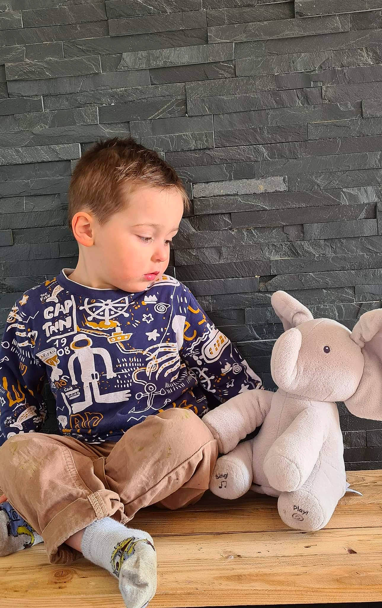 Owen participe au concours pour gagner de l'argent avec cette photo : baby_toddler_clothing, brick, brickwork, child, face, flooring, foot, happy, human_body, leg, pattern, person, plush, sitting, sleeve, sock, stuffed_toy, t_shirt, toddler, toy