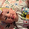Stella is registered to the contest to win money with this photo: baby, infant, bow, headwear, tongue, playful, cute, white_shirt, pineapple_design, colorful, polka_dot, floral_blanket, lying_down, child, portrait, close_up, skin, face, happy, adorable
