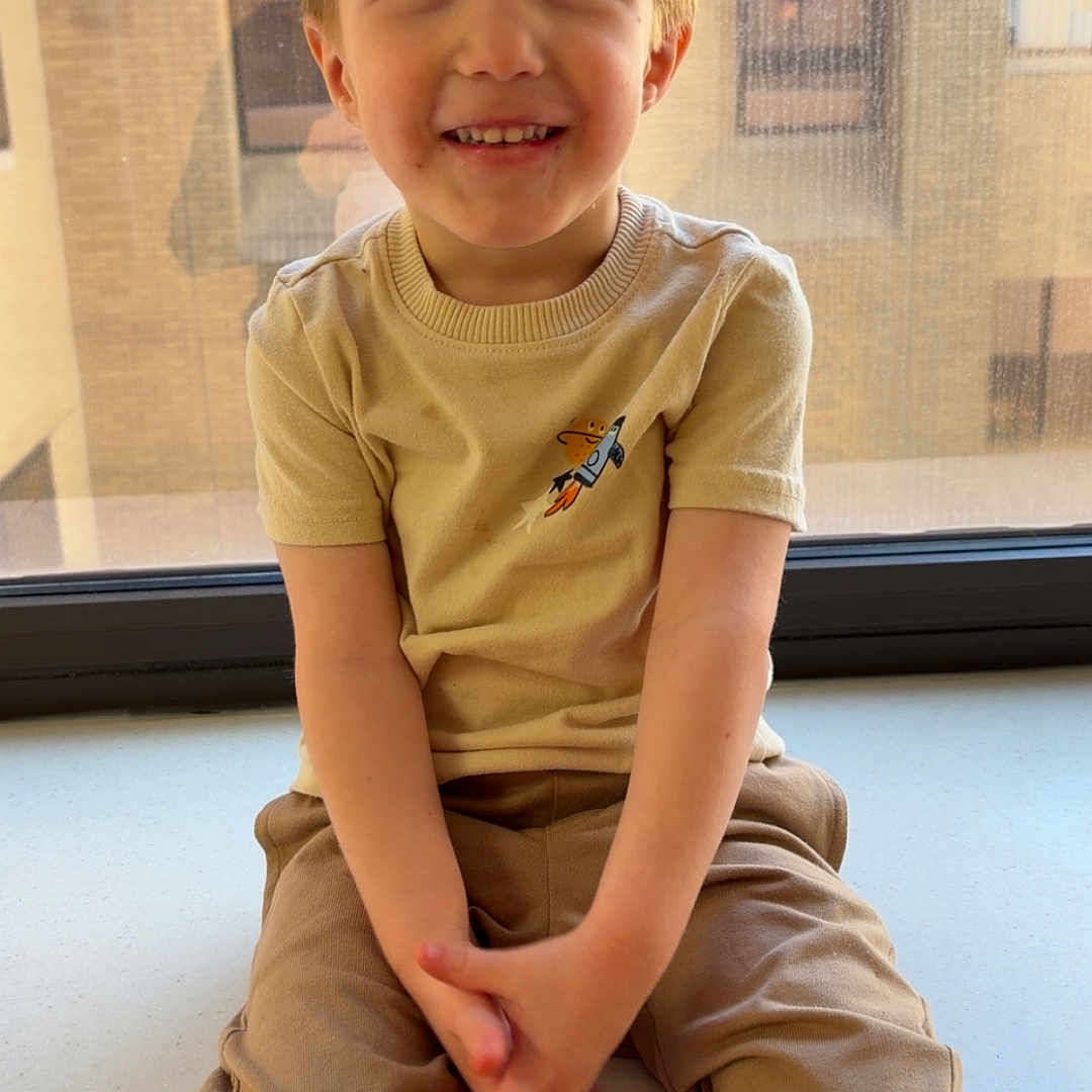 Leo is registered to the contest to win money with this photo: child, boy, smiling, window, windowsill, beige_clothing, short_sleeves, pants, indoor, natural_light, face, portrait, happy, sitting, casual, young, urban_background, window_screen, hands, expression