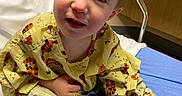 Leo is registered to the contest to win money with this photo: toddler, child, hospital, bed, gown, yellow, blue, socks, face, smile, indoor, medical, furniture, curly_hair, person, young_child, cute, healthcare, room, sitting