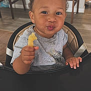 Brave is registered to the contest to win money with this photo: baby, child, high_chair, eating, snack, food, hand, smiling, pouting, portrait, indoor, dining_area, chair, table, wood_floor, home, gray_shirt, curly_hair, toddler, photography